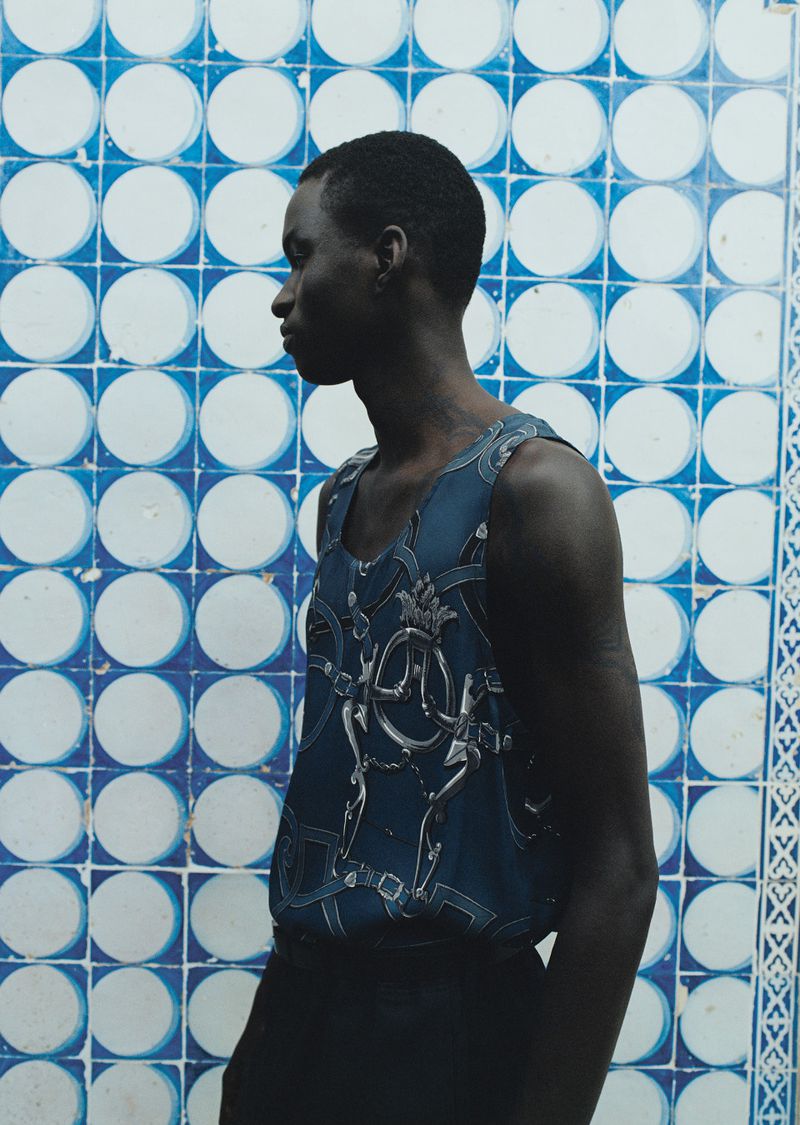 Profile portrait of an African American man wearing a patterned tank top against a blue and white patterned tile wall background.