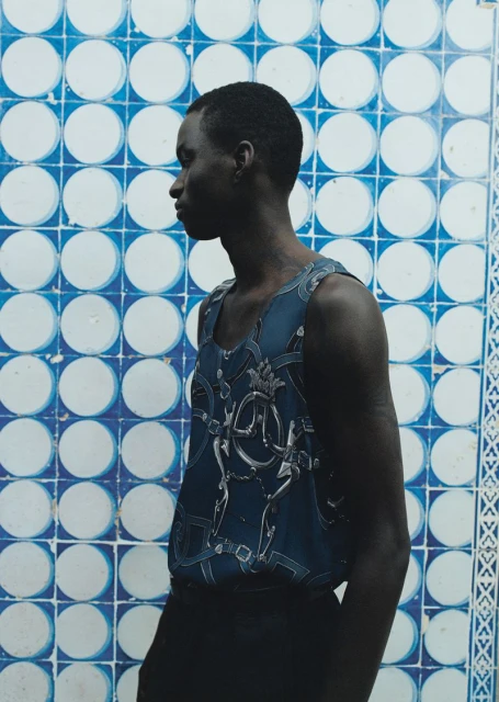 Profile portrait of an African American man wearing a patterned tank top against a blue and white patterned tile wall background.