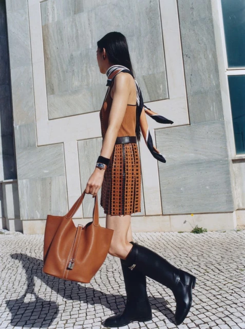 A stylish woman walks outdoors, wearing a brown dress, black boots, and carrying a tan tote bag on a sunny day.