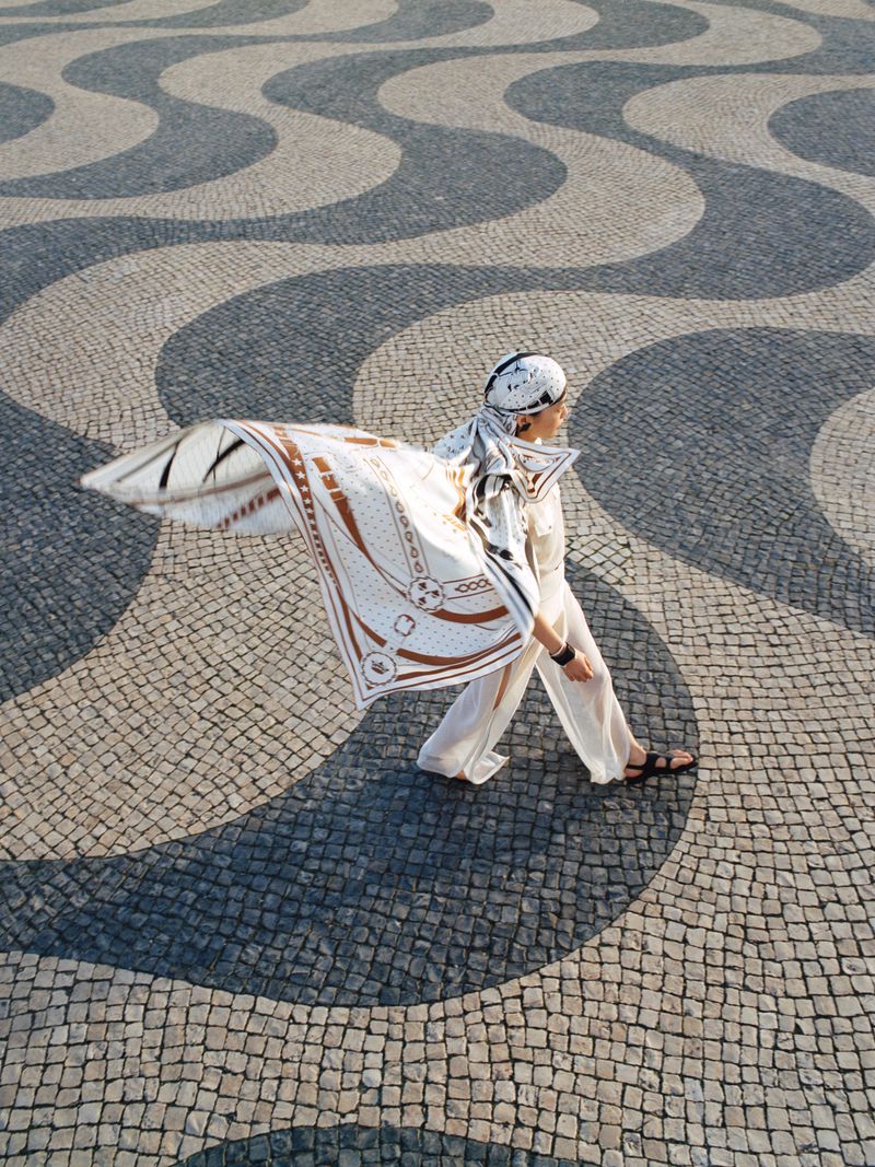 Woman in white walking across patterned cobblestone plaza wearing a patterned scarf and sandals in an outdoor environment.