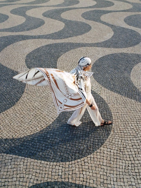 Woman in white walking across patterned cobblestone plaza wearing a patterned scarf and sandals in an outdoor environment.