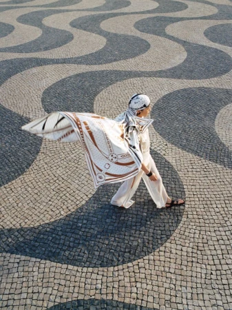 Woman in white walking across patterned cobblestone plaza wearing a patterned scarf and sandals in an outdoor environment.