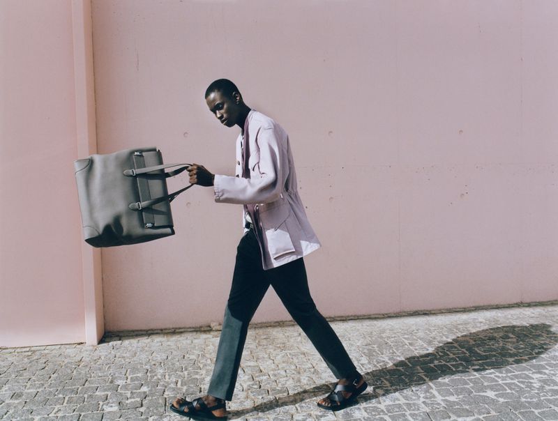 African American man in light jacket walking with grey duffel bag in an outdoor fashion editorial with a light pink wall backdrop.