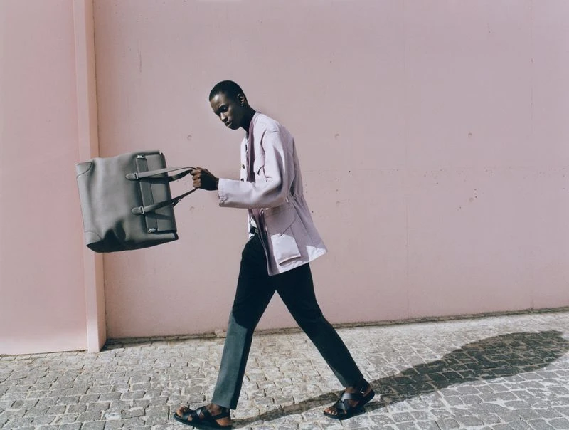 African American man in light jacket walking with grey duffel bag in an outdoor fashion editorial with a light pink wall backdrop.