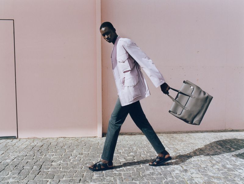 Stylish adult Black male strides along a cobblestone street, carrying a grey tote bag, with a pink wall in the background.