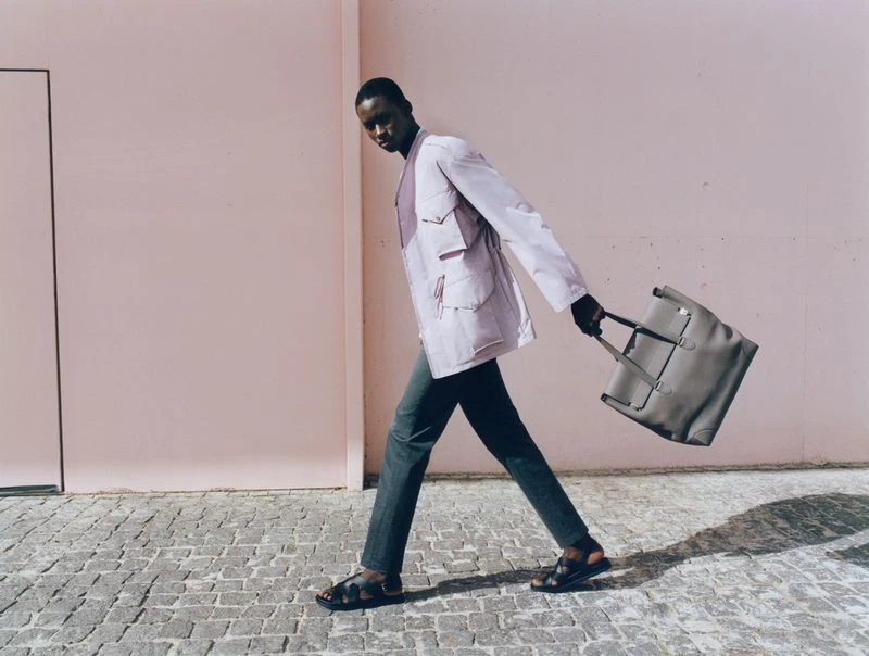 Stylish adult Black male strides along a cobblestone street, carrying a grey tote bag, with a pink wall in the background.