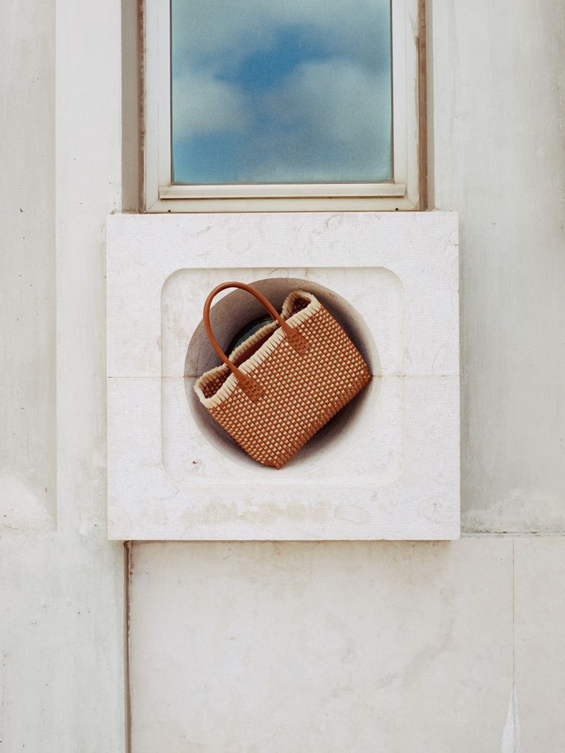 Woven handbag with brown leather handles inside white concrete circle, window with cloud reflection on the top in daylight.