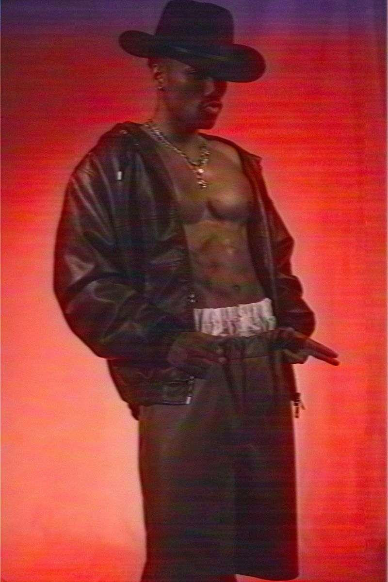 A Black male fashion model strikes a pose in a studio against a red backdrop, wearing a black cowboy hat, leather jacket, and skirt.
