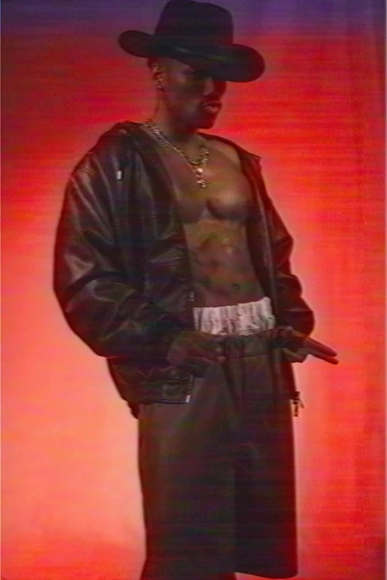 A Black male fashion model strikes a pose in a studio against a red backdrop, wearing a black cowboy hat, leather jacket, and skirt.