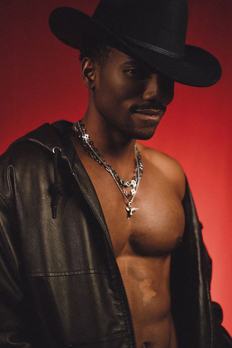 Black man wearing a black cowboy hat and leather jacket with layered necklaces against a red background