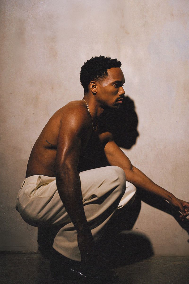 Portrait of an adult African-American male crouching indoors; he wears light pants and a chain, photographed in warm lighting.