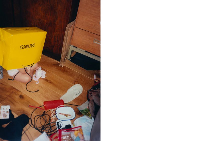 A child is partially visible behind a large yellow Selfridges shopping bag on a wood floor surrounded by various objects and clutter.