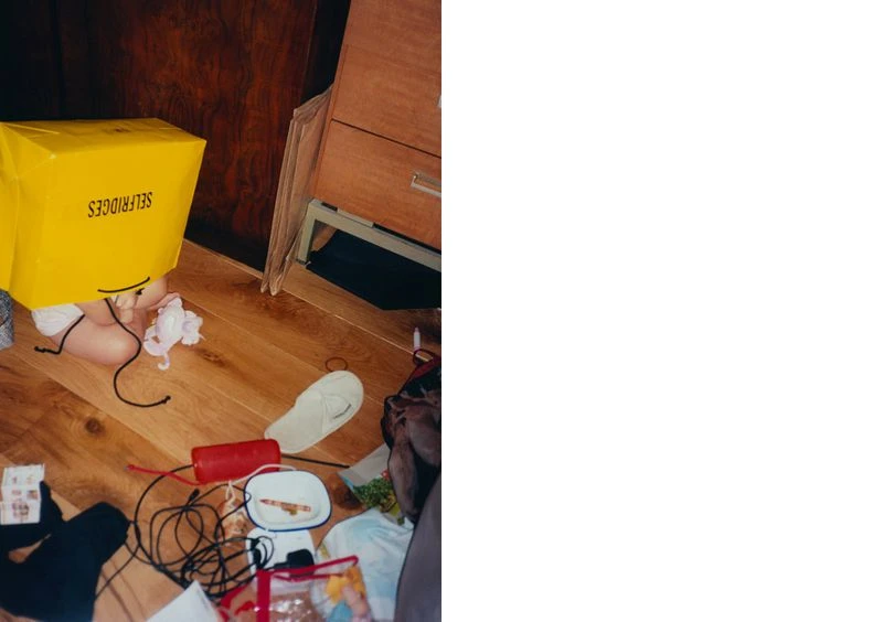 A child is partially visible behind a large yellow Selfridges shopping bag on a wood floor surrounded by various objects and clutter.