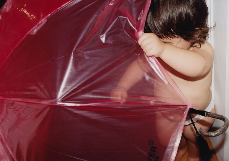 Infant peers from behind a translucent pink umbrella indoors, revealing dark hair and partial view of skin in a candid shot.