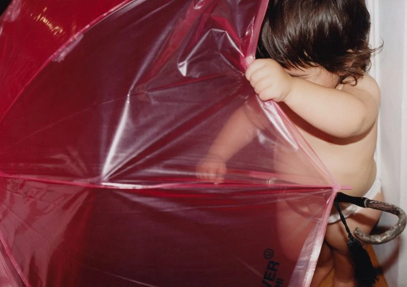 Infant peers from behind a translucent pink umbrella indoors, revealing dark hair and partial view of skin in a candid shot.
