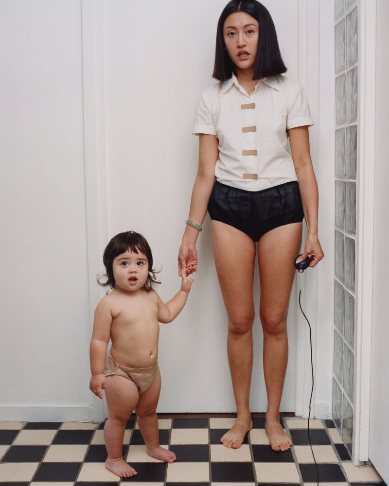 A woman and a toddler hold hands and look directly at the camera in an indoor studio setting with a checkered floor and neutral colors.