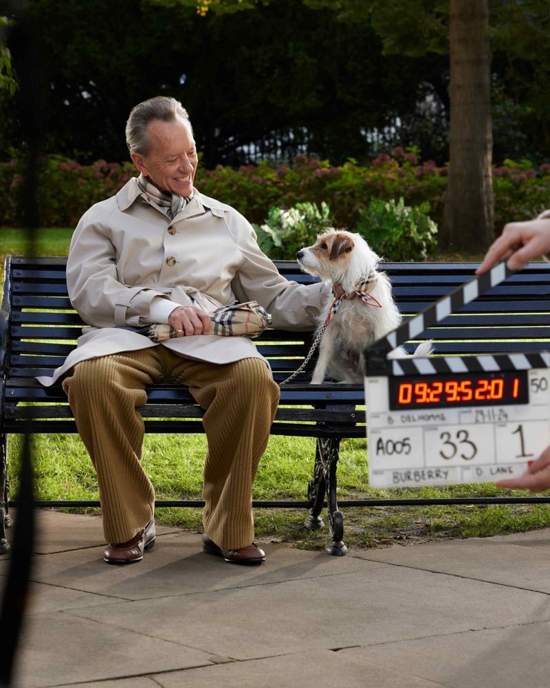 Elderly man sits on a bench with his dog during a Burberry fashion shoot, featuring a clapperboard, in an outdoor park setting.
