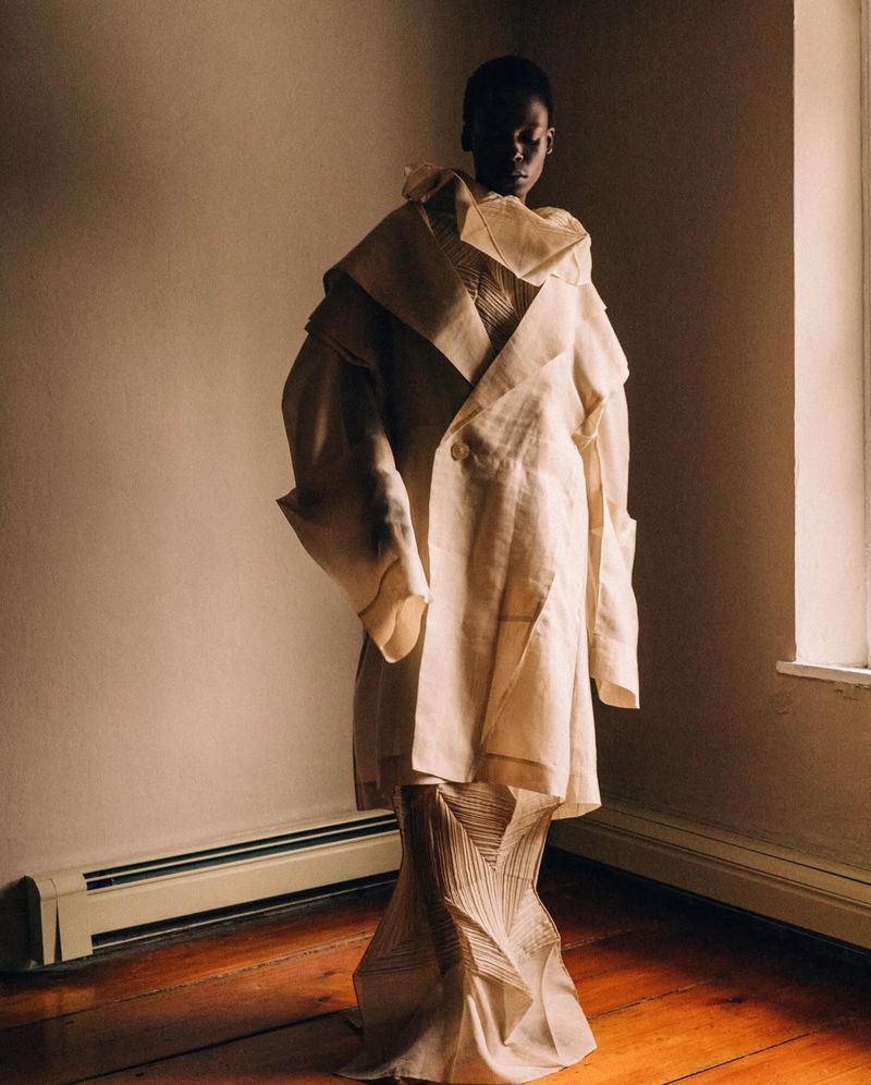 An adult female stands indoors with a neutral expression, wearing a textured, layered beige avant-garde dress in front of a heater and window.