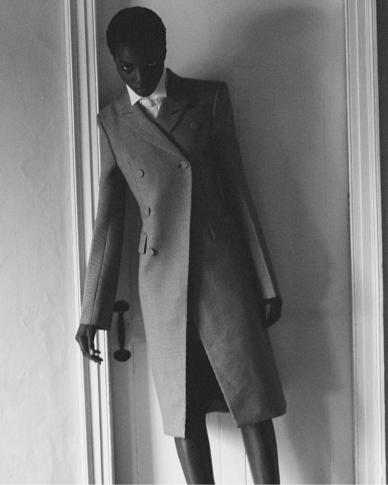 A statuesque Black woman in a grey coat, white shirt, and black skirt poses in a doorway, captured in elegant black and white.