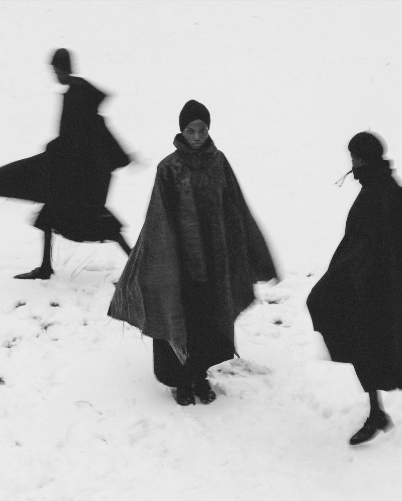 Black-and-white fashion shot with three figures in dark capes moving across a snowy field, one model centered and facing the camera.