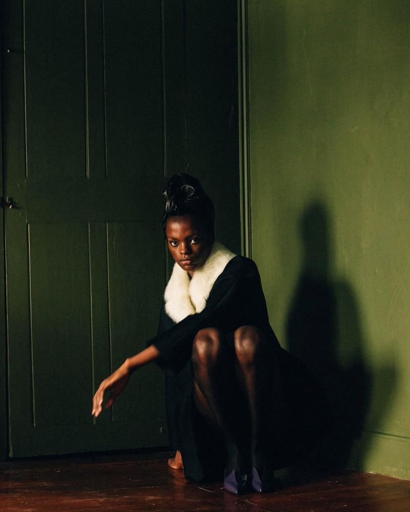 Fashionable Black woman squats indoors in a black wool coat with white fur collar, wearing sheer stockings and blue high heel shoes.