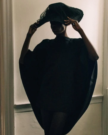 An African American woman wears a dramatic black hat and flowing poncho indoors against a light-colored wall for a fashion editorial shoot.