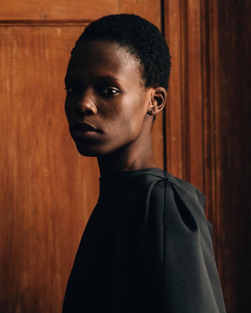 Close-up portrait of a young African-American woman in a draped black dress against a warm brown wooden paneled background indoors.