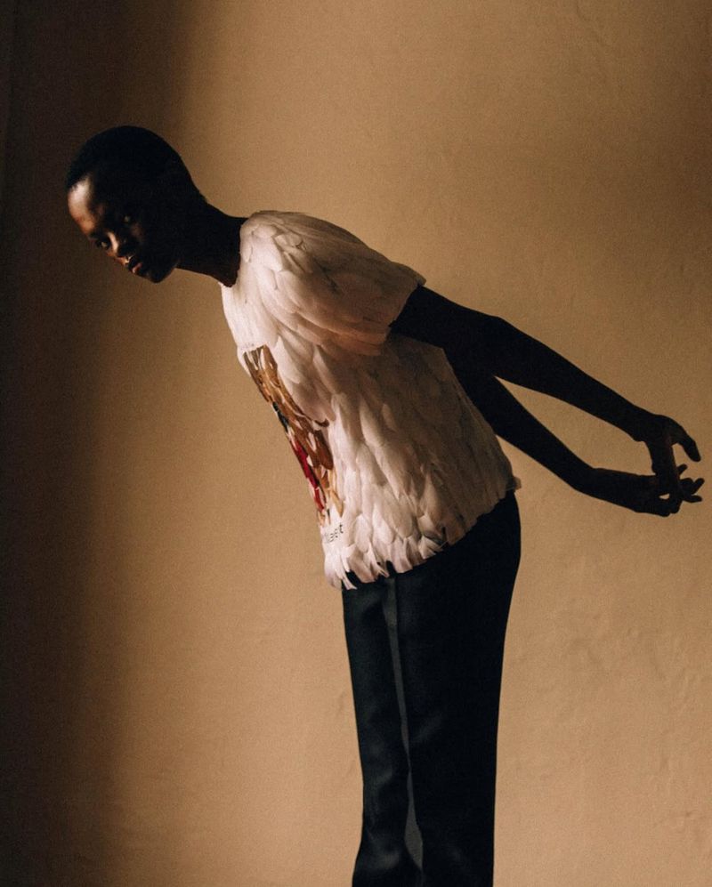 Elegant studio portrait of an African-American man in a feather-trimmed T-shirt and black pants, bending to the side, against a neutral backdrop.