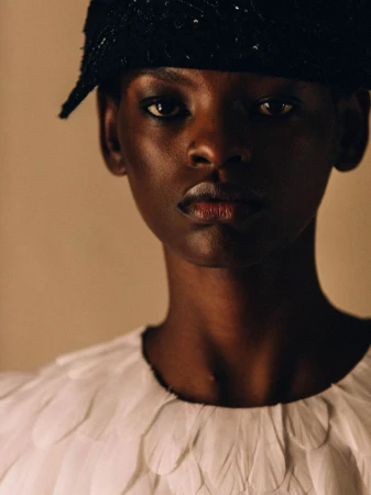 Close-up portrait of a model wearing a dark embellished hat and a white textured top against a neutral background.