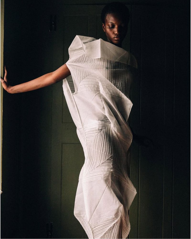Elegant, dark-skinned woman poses in a sculptural white dress with folded fabric details against a dimly lit, dark green doorway.