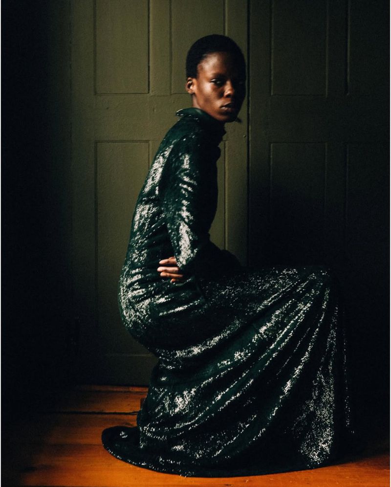 A single, slim African woman poses indoors in a long-sleeved, dark-green, shimmering gown, looking at the camera in a dimly lit setting.