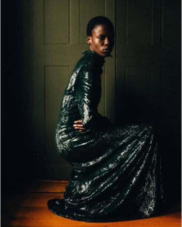 A single, slim African woman poses indoors in a long-sleeved, dark-green, shimmering gown, looking at the camera in a dimly lit setting.