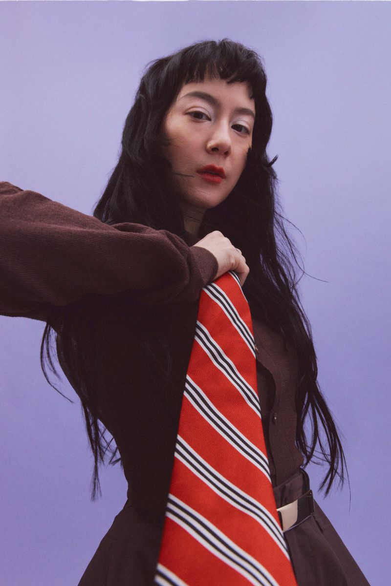 Portrait of an adult woman in a brown dress holding a red striped necktie against a light purple backdrop in a studio setting.
