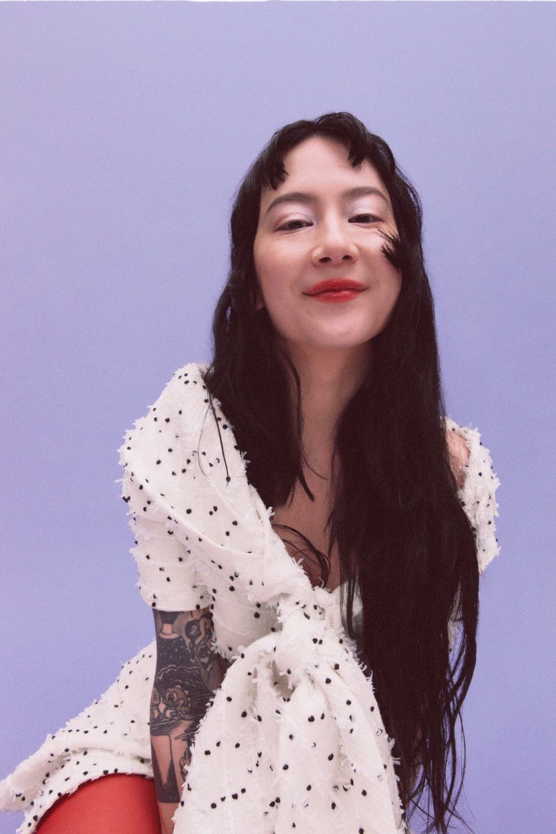 Close-up of a woman with long dark hair wearing a white dress with black dots, posing against a solid light purple backdrop.