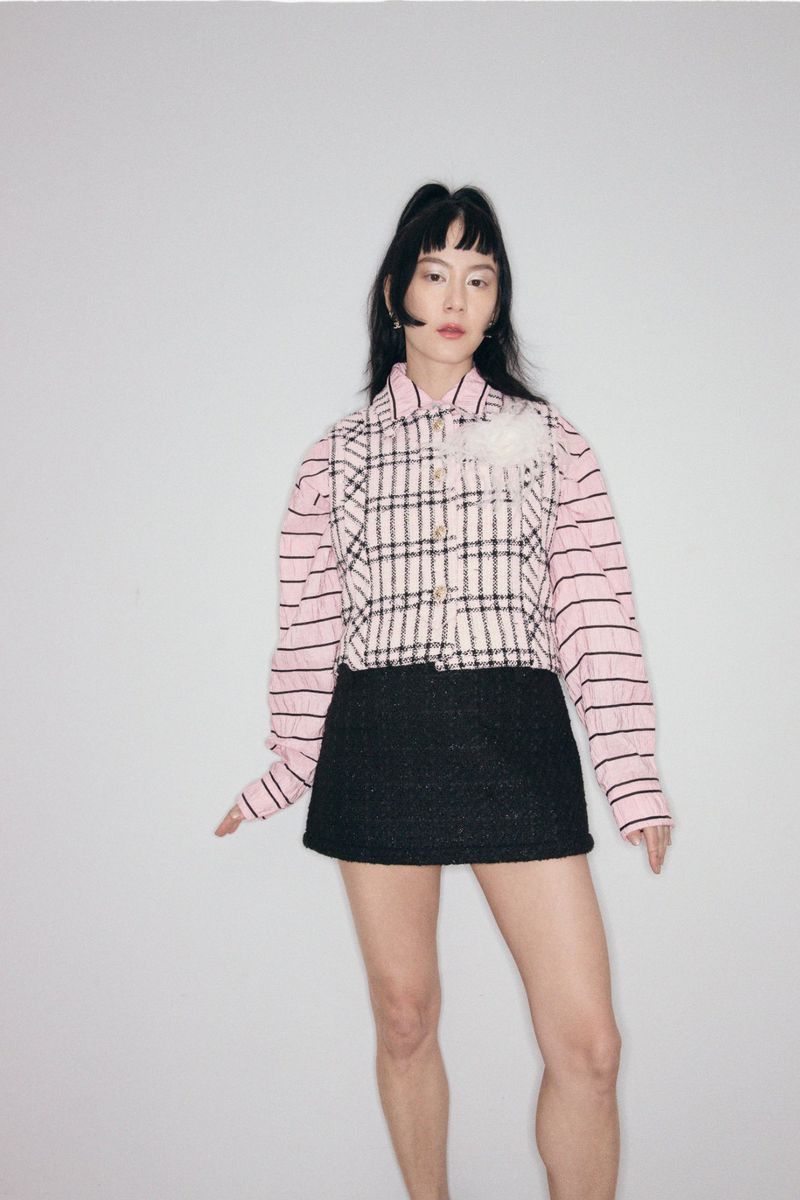 Adult woman posing in a pink-and-white plaid jacket, black skirt, and jewelry stands against a plain white studio backdrop.