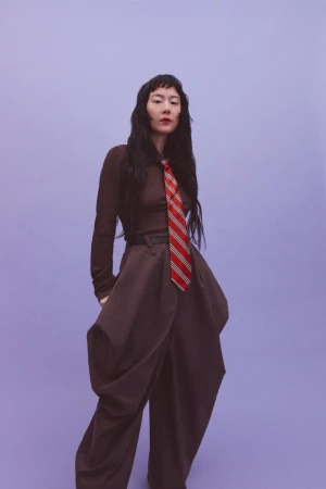 Woman with long dark hair wearing a brown outfit and striped tie against a purple background