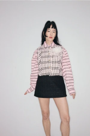 East Asian woman wearing a pink striped shirt, plaid vest, and black skirt against a grey seamless background