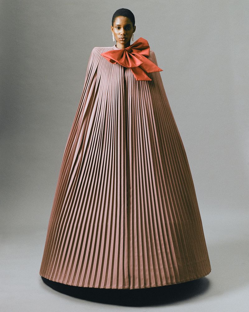 A solo African-American female fashion model poses in a light studio backdrop wearing a floor-length pleated cape-dress with large bow.