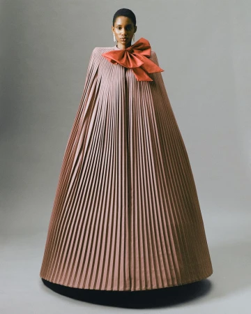 A solo African-American female fashion model poses in a light studio backdrop wearing a floor-length pleated cape-dress with large bow.