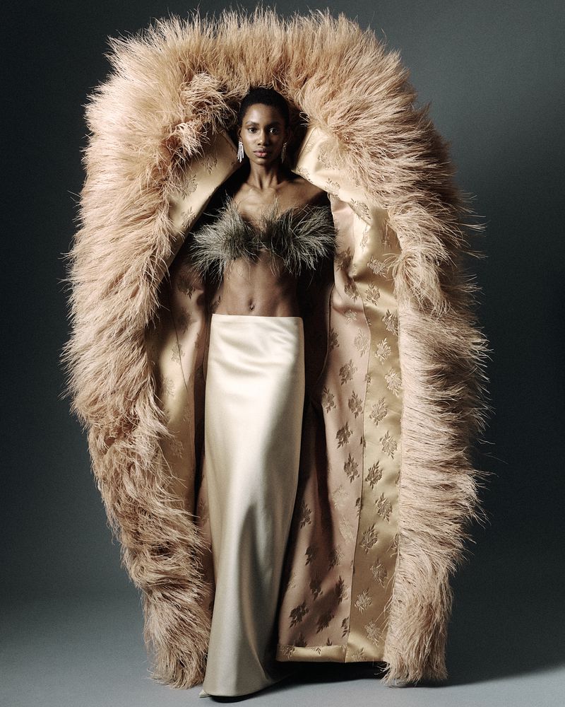 Fashion portrait of an adult woman in a faux fur coat with a long skirt, wearing dangling earrings, against a grey backdrop.