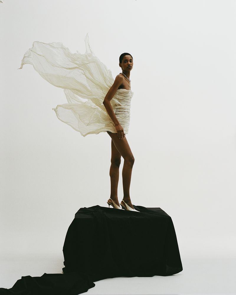 Young woman in a white dress stands on a black surface, posing in front of a light background in a fashion style studio shot.
