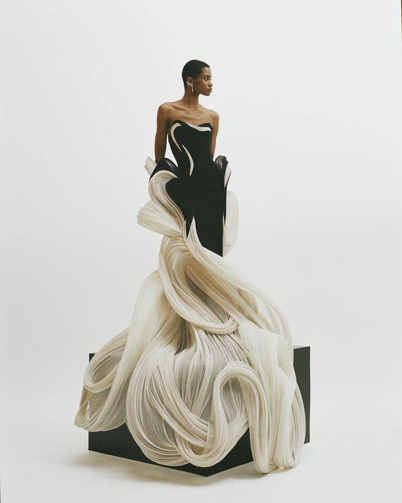Model in dramatic black and white gown with swirling fabric detail, posing against a white backdrop in a fashion forward photo shoot.