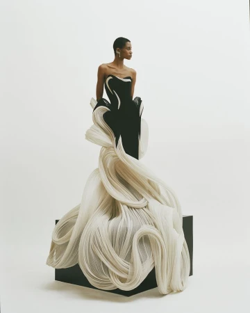 Model in dramatic black and white gown with swirling fabric detail, posing against a white backdrop in a fashion forward photo shoot.
