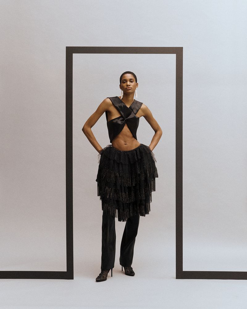 A slender Black woman poses in avant-garde fashion, featuring a twisted top and layered tulle skirt, framed by a dark border on a white backdrop.