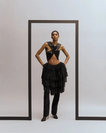 A slender Black woman poses in avant-garde fashion, featuring a twisted top and layered tulle skirt, framed by a dark border on a white backdrop.
