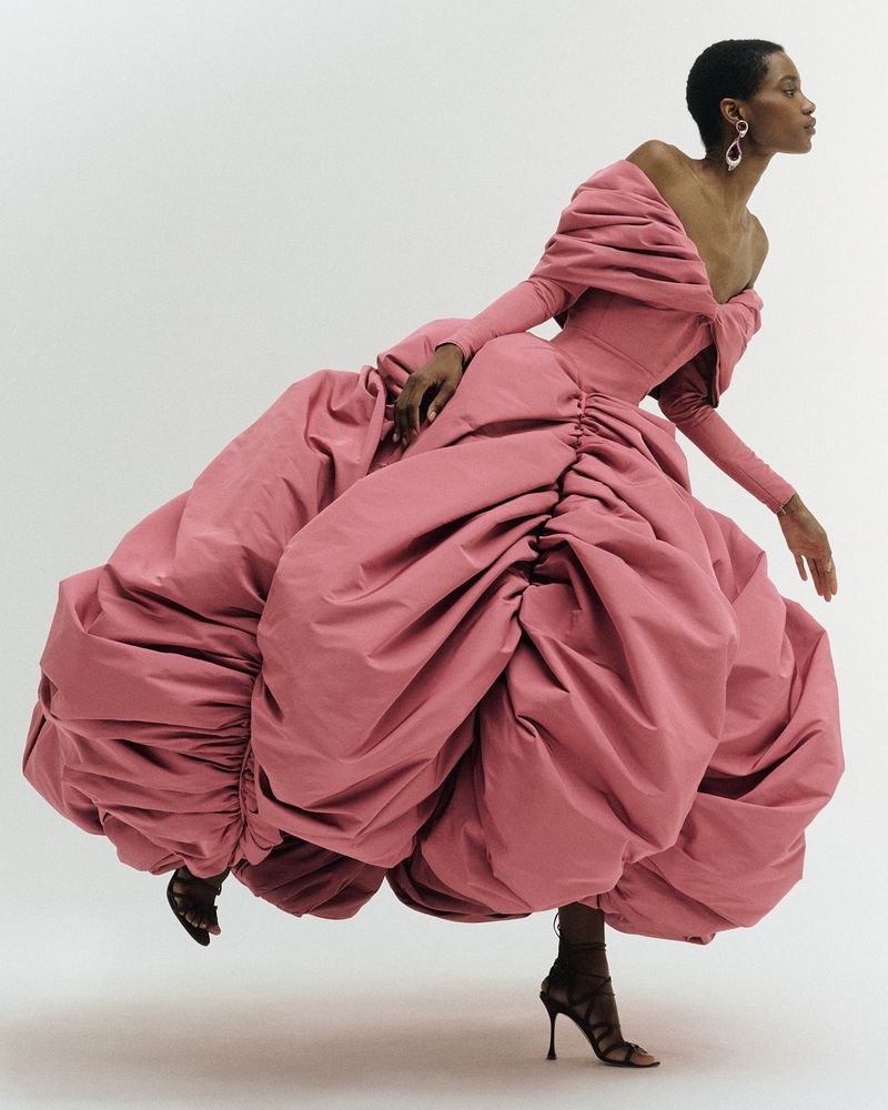 A fashion model poses in a dramatic, pink, off-the-shoulder gown with a large, voluminous skirt. The model wears strappy heels in a studio setting.