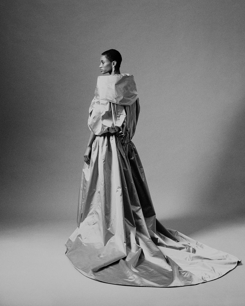 A full, side view of a Black female fashion model in a silver, high-fashion gown with a dramatic collar posing in a photo studio.