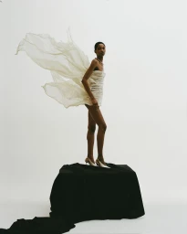 Model standing on a draped black pedestal in a white studio wearing a white dress with flowing fabric wings.
