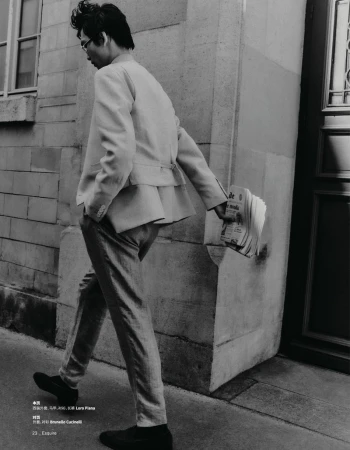 Stylish man walks in light suit, carrying newspaper. Classic fashion, neutral tones, architectural backdrop, and editorial aesthetic.