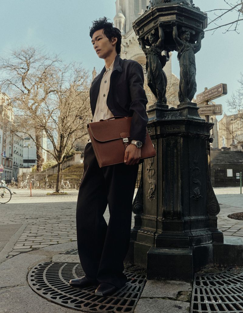Asian man in black suit with brown leather briefcase poses near ornate streetlamp in European city against a blue sky background.